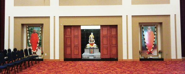 A large stone image Shiva is located off the far side of the auditorium, behind sliding panels, flanked by large framed pictures of Swami Tapovan Maharaj (left) and Swami Chinmayananda (right).